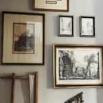 A wall with various framed artworks, including sketches and prints, arranged around a wooden ladder leaning against the wall.